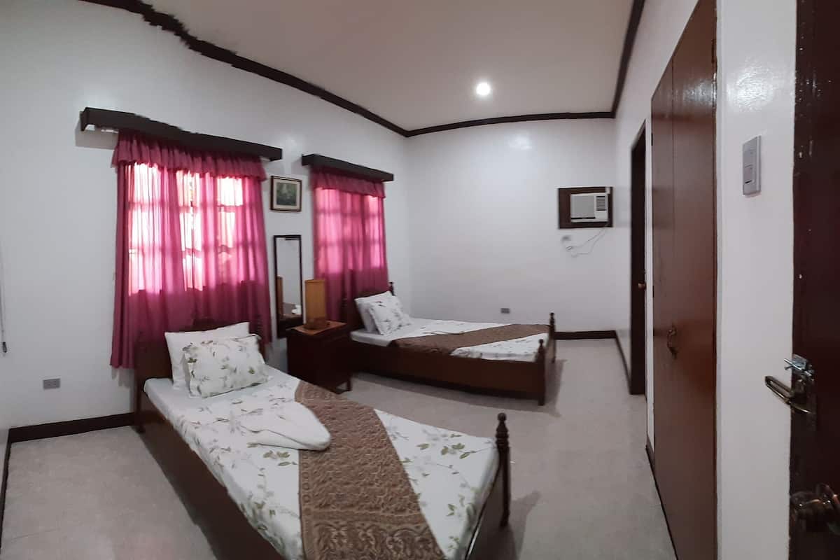 Quarto com duas camas de solteiro, uma pequena mesa de cabeceira, um criado-mudo, um ar condicionado fixado na parede e uma porta com uma placa.