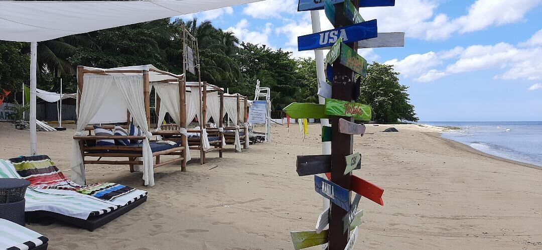 A beach with wooden beds under a canopy, a signpost with multiple directional signs, and a sandy beach with palm trees in the background.