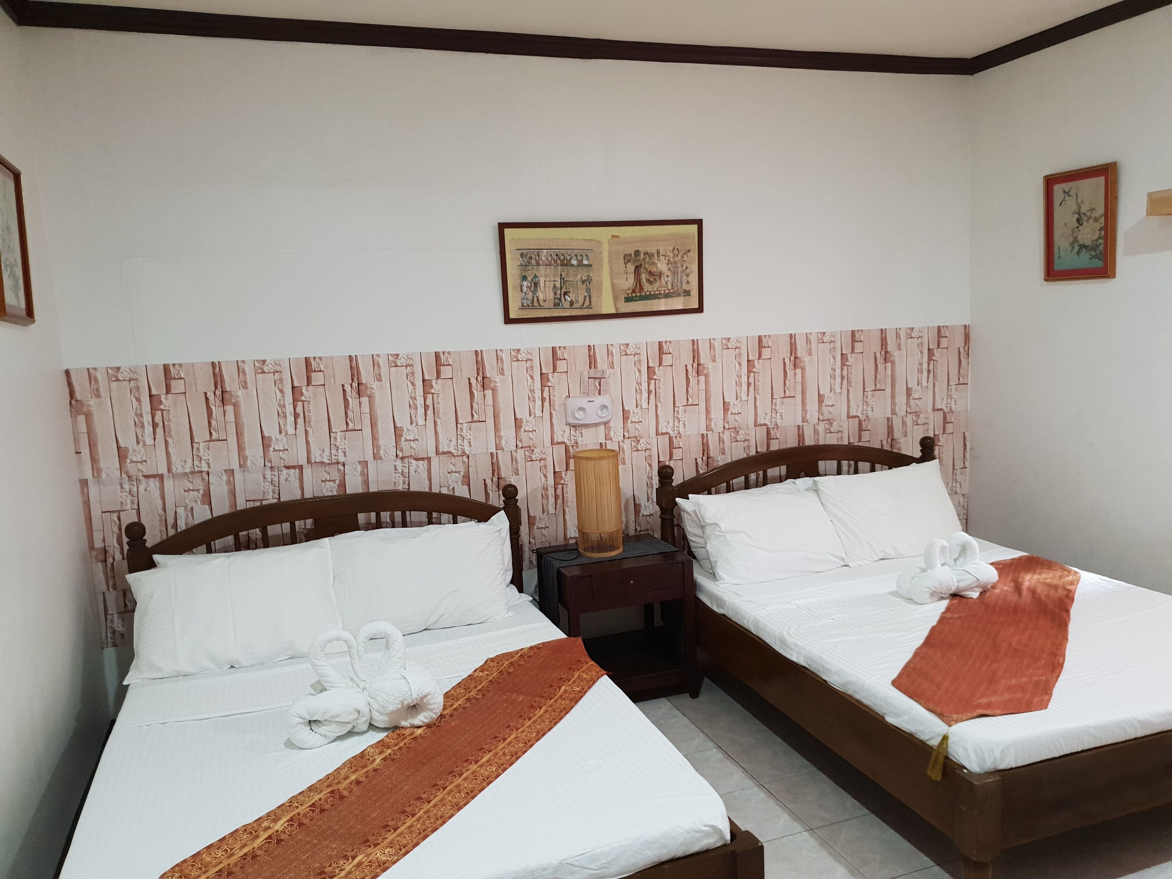 Una habitación de hotel con dos camas, cada una con ropa blanca y cabeceros marrones. Hay una lámpara de pared en la mesita de noche junto a una cama, y un cuadro enmarcado sobre el cabecero.