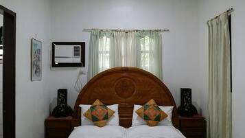 A bedroom with a wooden headboard, two bedside tables, a bed with white bedding, a wall-mounted air conditioner, and a window with sheer curtains.
