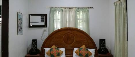 A bedroom with a wooden headboard, two bedside tables, a bed with white bedding, a wall-mounted air conditioner, and a window with sheer curtains.