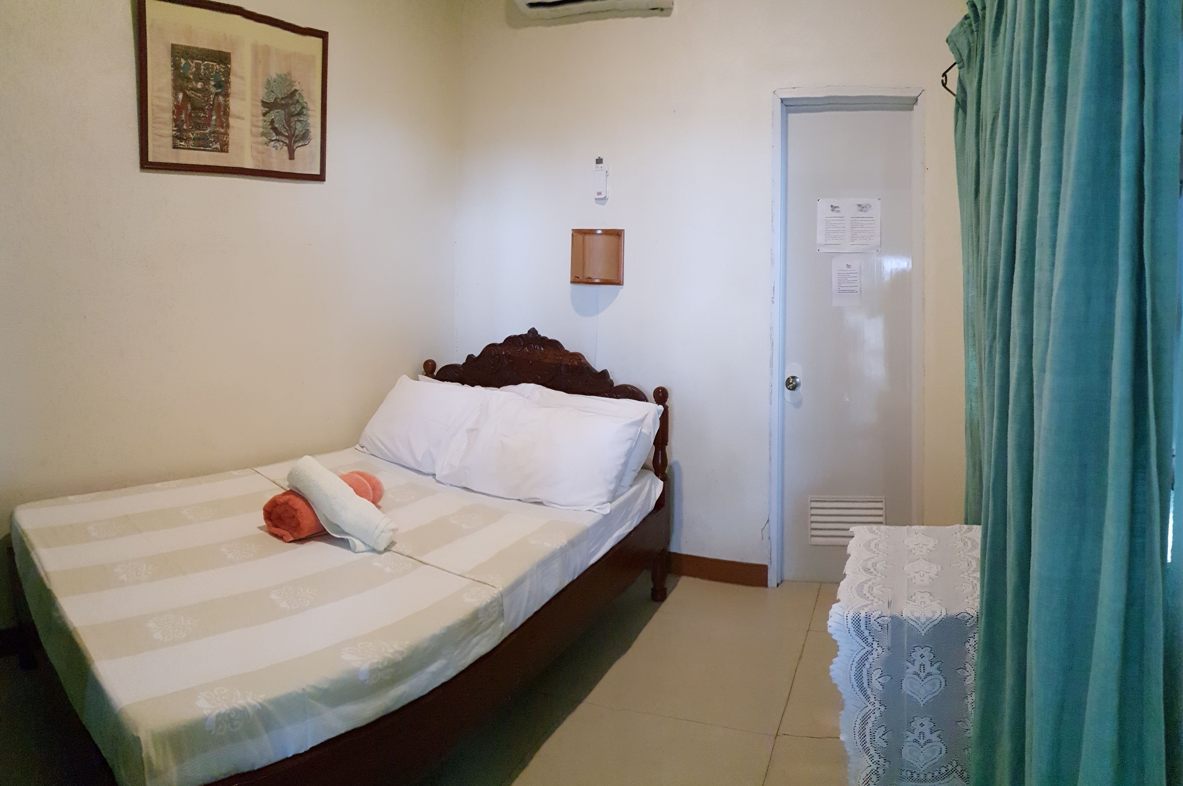 A single bed with a striped bedspread, a wooden headboard, and a white pillow. There is a framed picture on the wall, a door with a white curtain, and a small wooden shelf with a light.