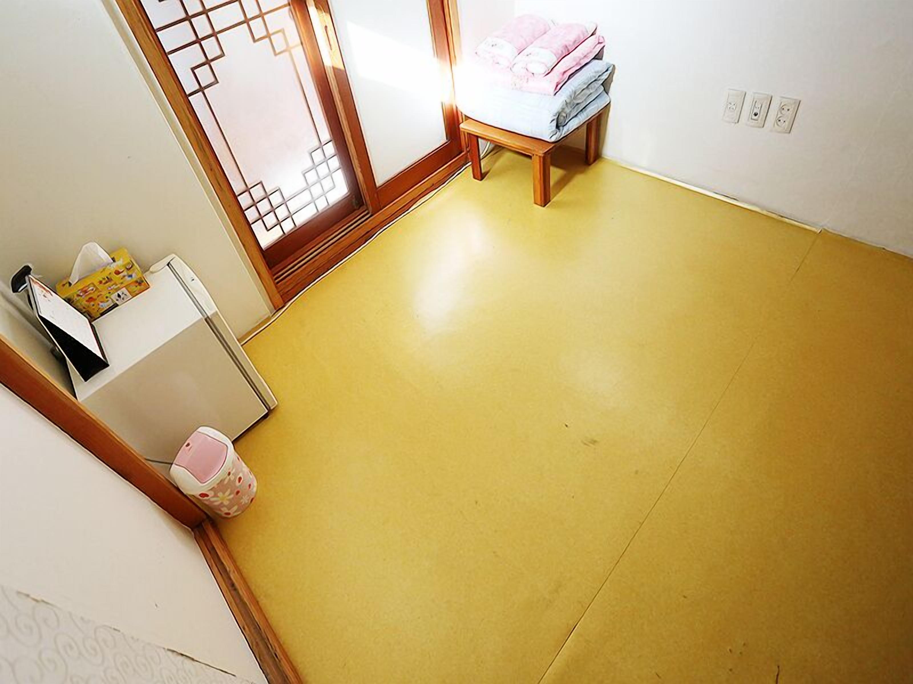 Una habitación con piso amarillo, una cama de madera pequeña con ropa de cama rosa, un armario blanco y una puerta corrediza de vidrio con un diseño geométrico.
