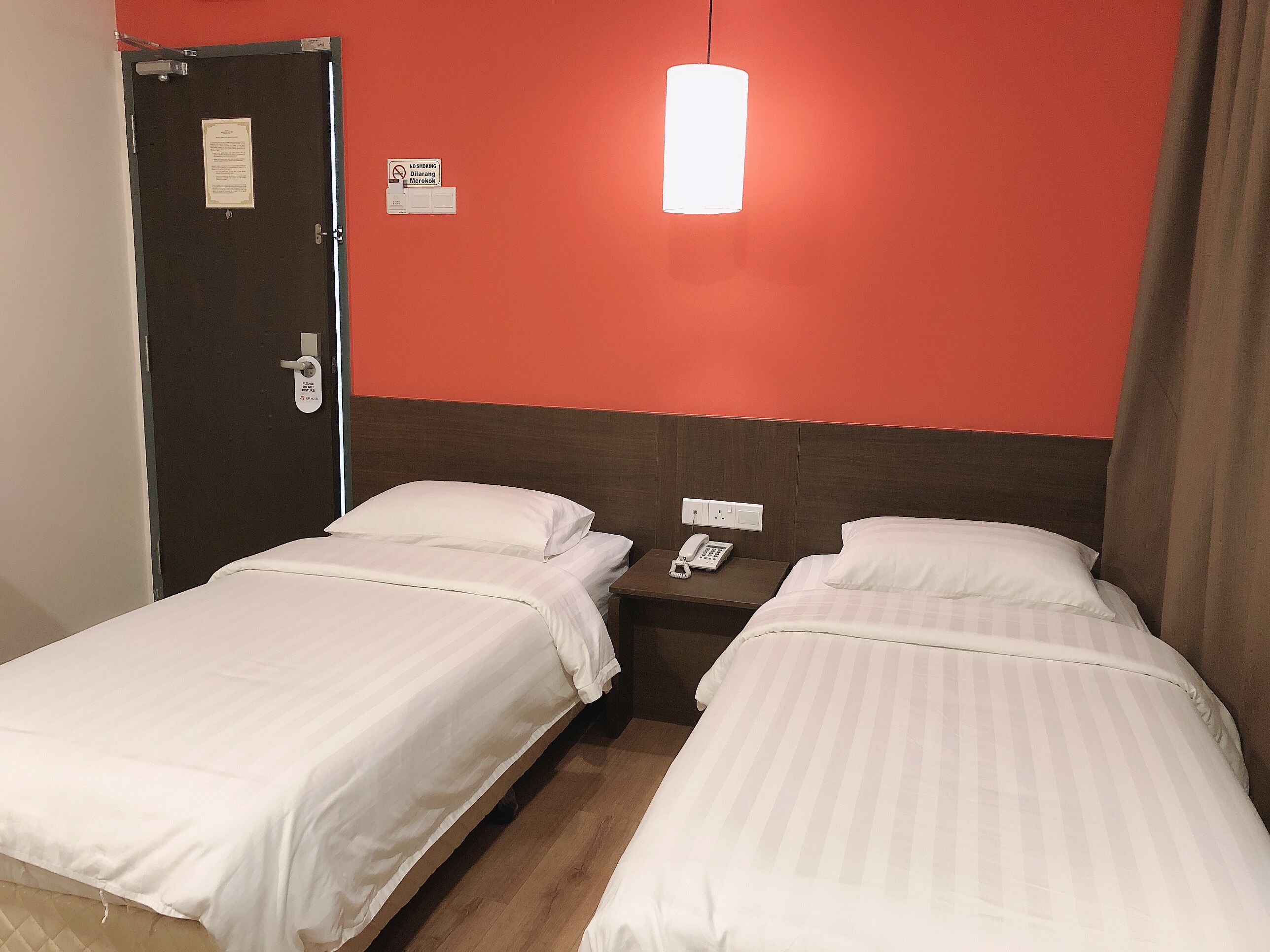 Habitación de hotel con dos camas, una mesita de noche con teléfono, pared roja y una puerta con un cartel.