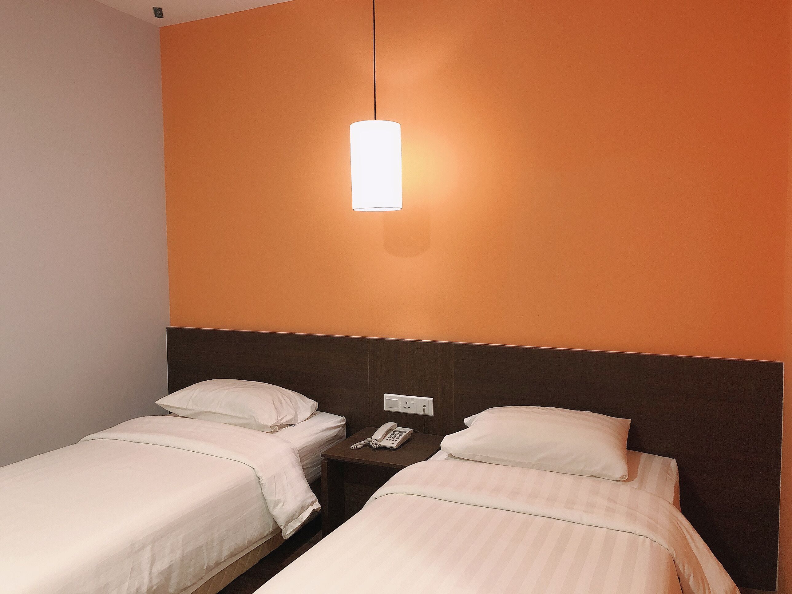 A hotel room with two single beds, a nightstand with a phone, and a wall-mounted light fixture.