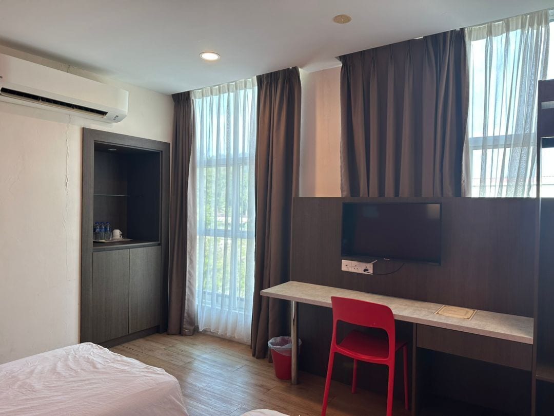 A hotel room with a bed, a desk with a chair, a TV, a wardrobe, and a window with curtains.