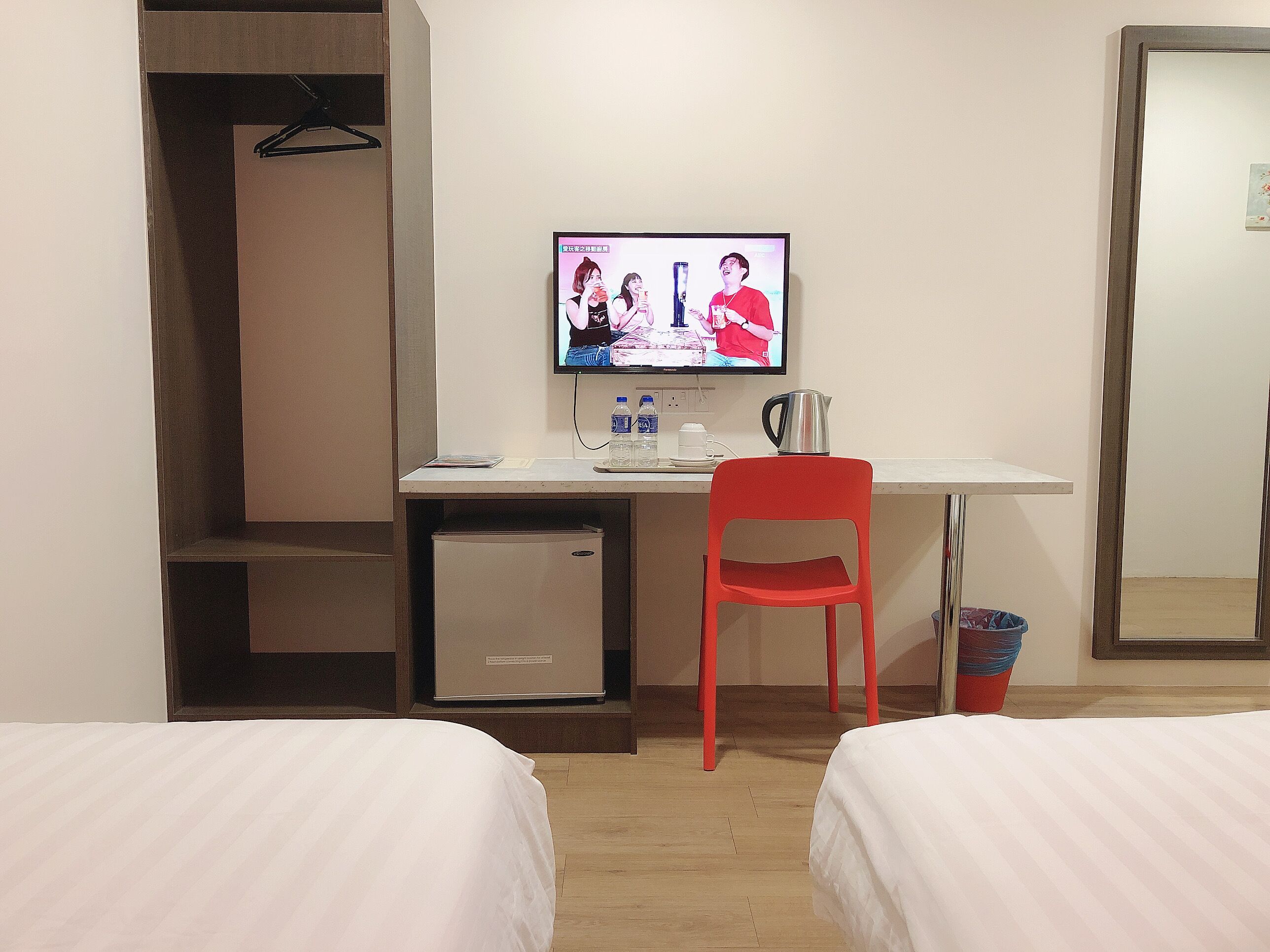 A hotel room with a bed, a desk with a computer, a red chair, a small fridge, and a mirror.