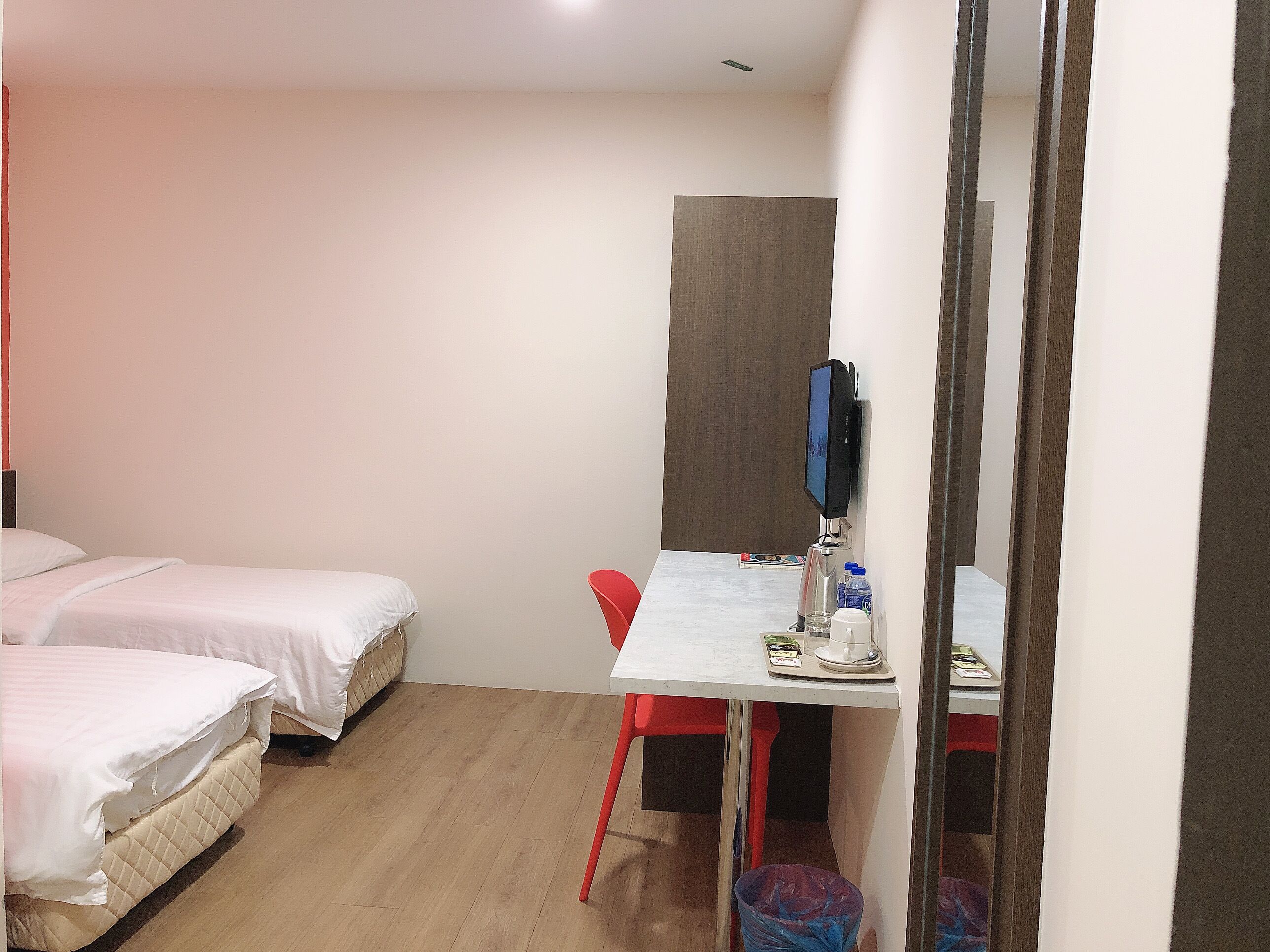 Une chambre d’hôtel avec deux lits, un bureau avec une chaise, une télévision et une poubelle.
