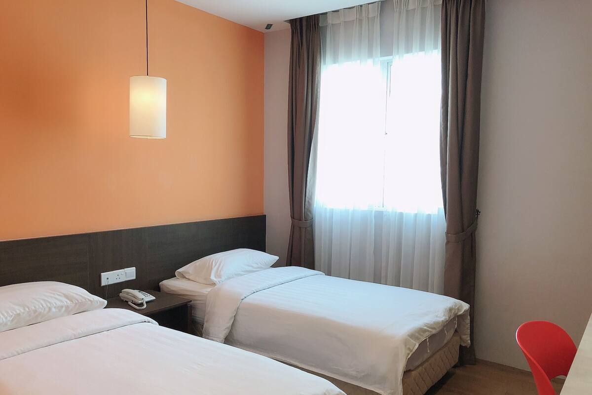 A hotel room with two beds, a bedside table with a phone, a lamp, and a window with curtains.