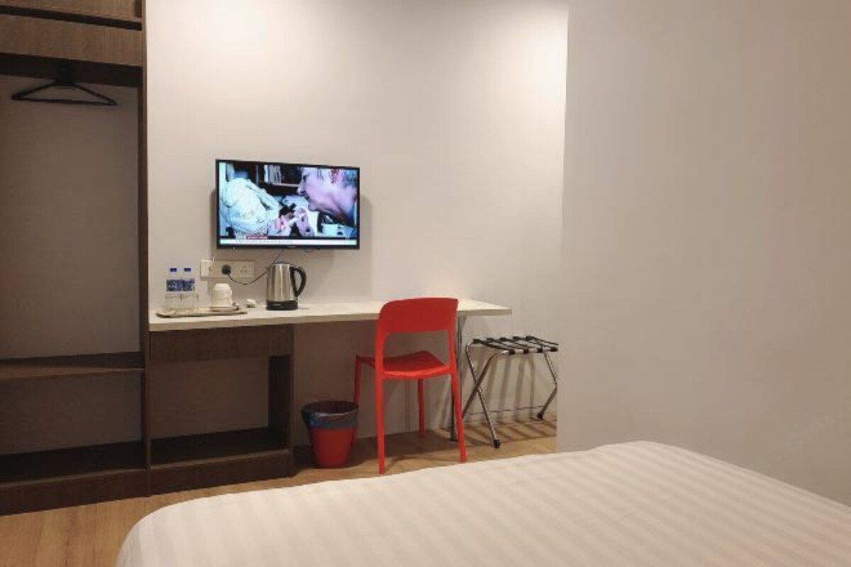 Ett hotellrum med en säng, ett skrivbord med en tv, en röd stol, en vattenkokare och en liten papperskorg.