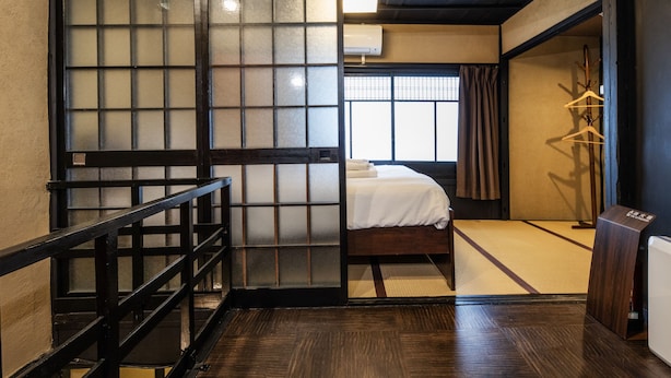 A traditional Japanese room with tatami flooring, a bed with white bedding, a sliding door, and a wooden shelf. - Shiki Homes NATSU (Kyoto)