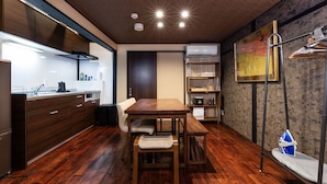 A modern kitchen with wooden cabinets, a dining table, and a bench. There is a built-in oven, a microwave, and a coffee machine. The room has a wooden floor and a dark ceiling with recessed lighting. - Shiki Homes NATSU (Kyoto)
