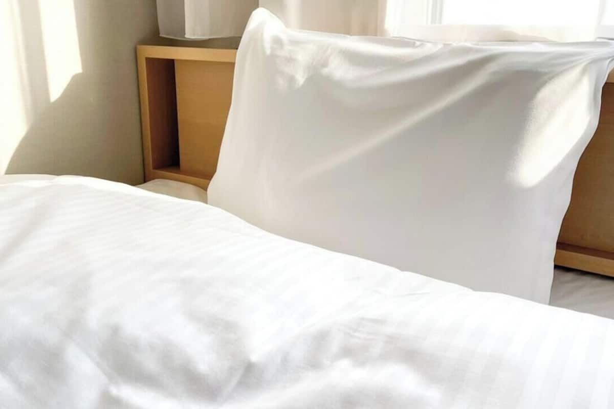 Huoneessa on sänky, jossa on valkoiset vuodevaatteet, sekä puinen yöpöytä ja valkoinen lamppu.
