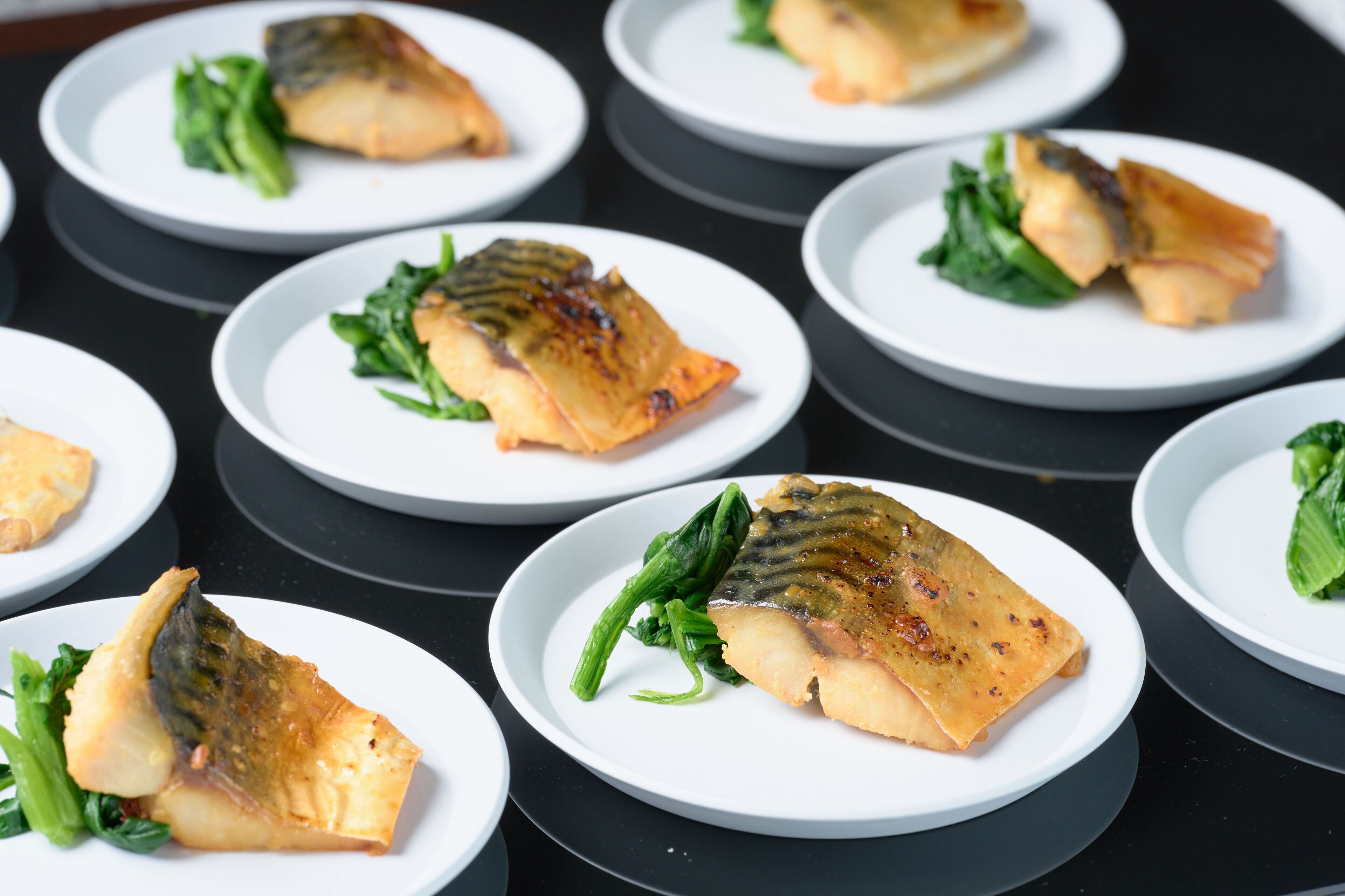 A series of white plates with food items, possibly fish, accompanied by greens.