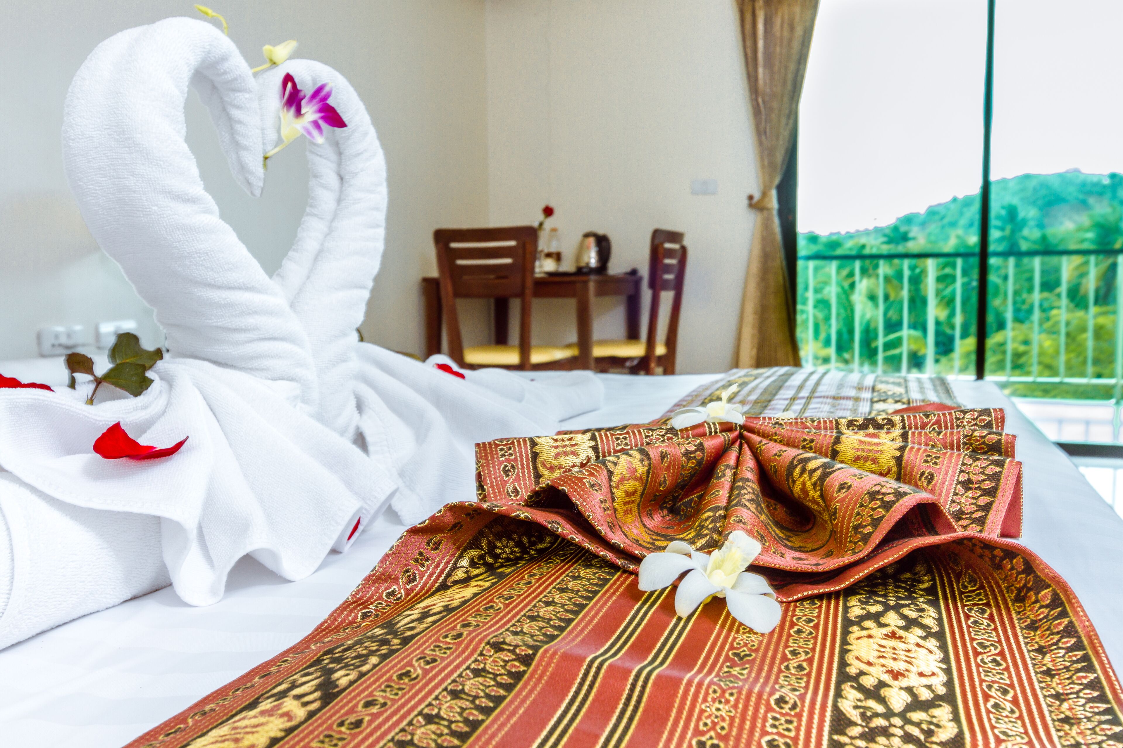 Una cama bien hecha con toallas blancas y un paño rojo estampado, una silla de madera, una mesita con un florero, y un balcón con vistas a la vegetación.
