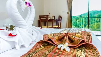 Una cama bien hecha con toallas blancas y un paño rojo estampado, una silla de madera, una mesita con un florero, y un balcón con vistas a la vegetación.