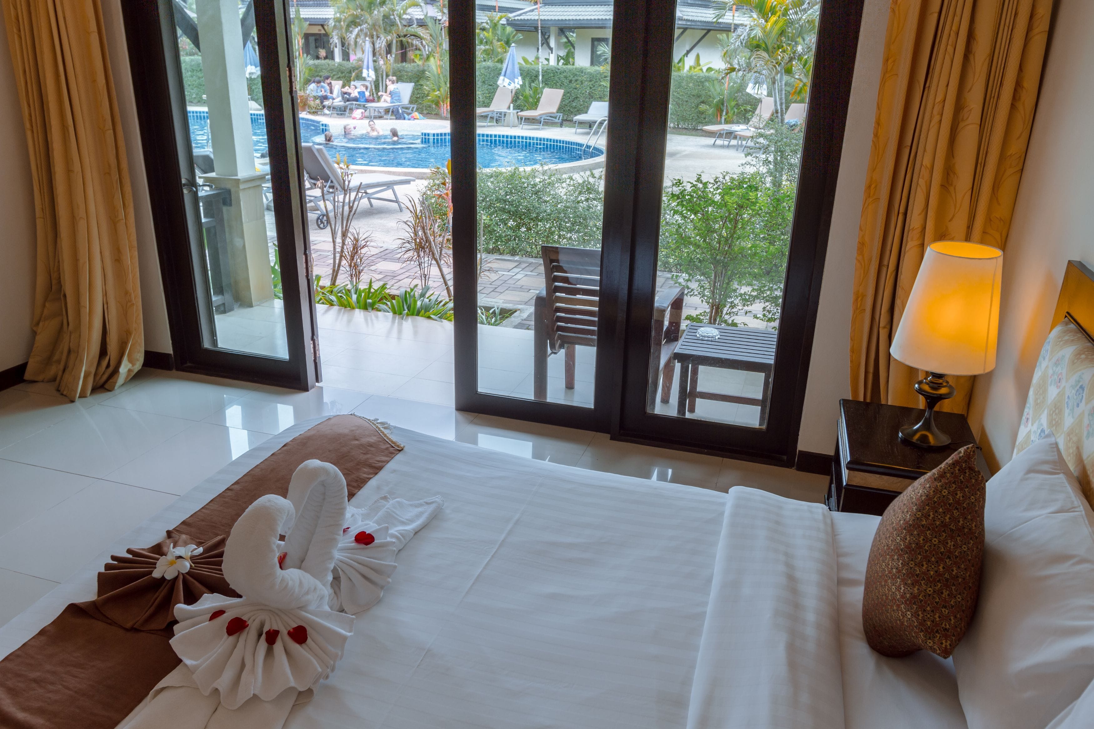 Ein Hotelzimmer mit Bett, Nachttisch mit Lampe und Blick auf Pool und Außenbereich mit Sitzgelegenheiten.