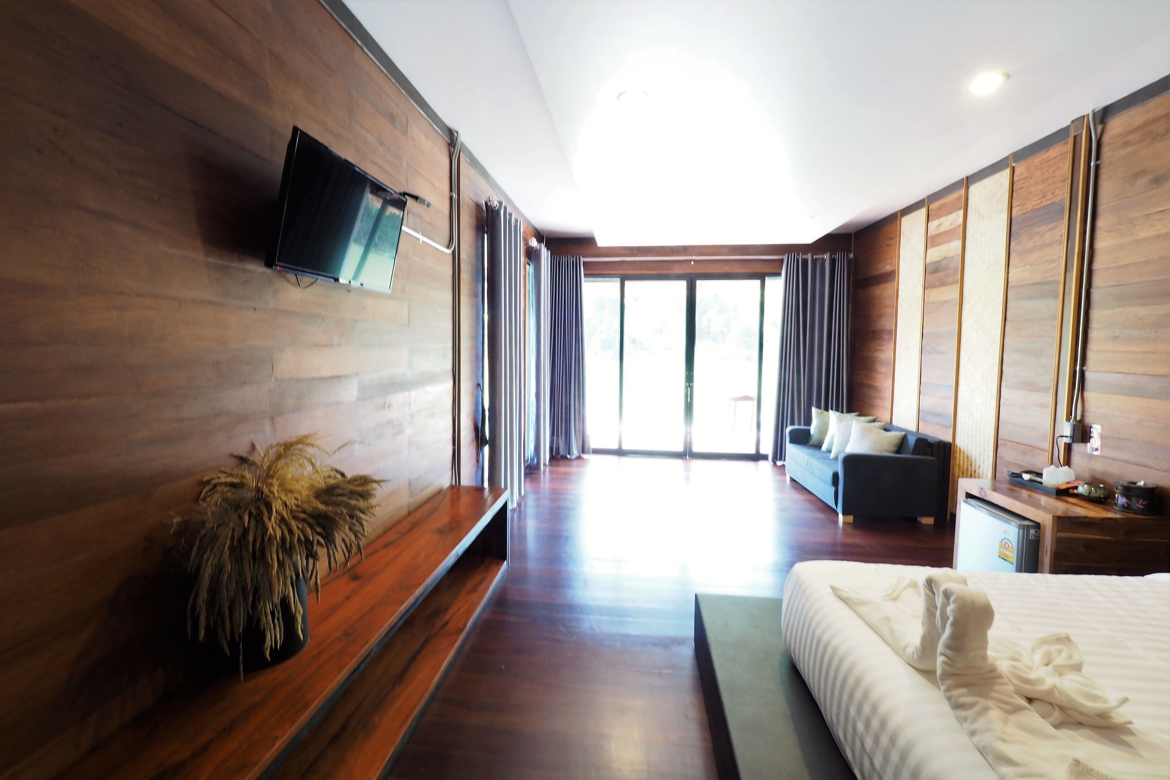 A modern hotel room with a wooden floor, a bed with white linens, a flat-screen TV mounted on the wall, a small desk with a lamp, and a large window with curtains.