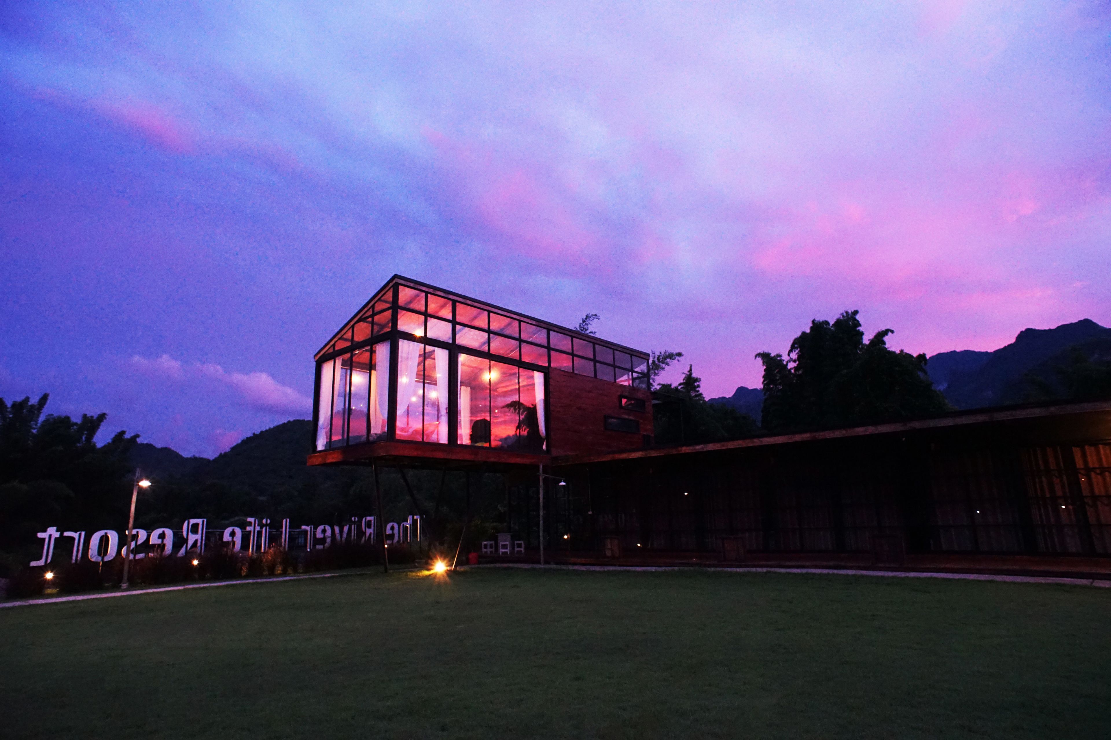A modern glass building with a flat roof, surrounded by a grassy area and mountains in the background.