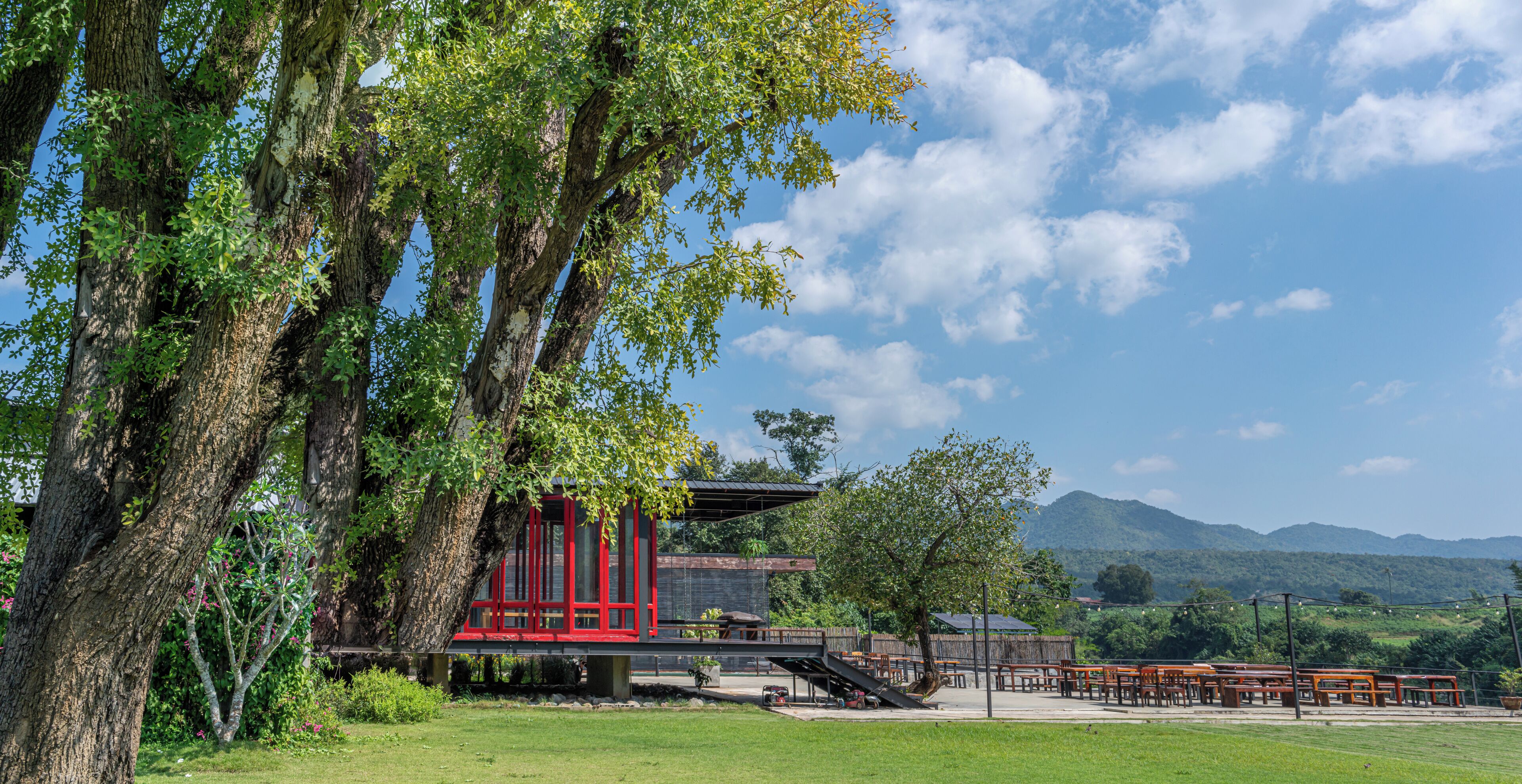 A red building with a covered outdoor area, surrounded by trees and a grassy field, with mountains in the background.