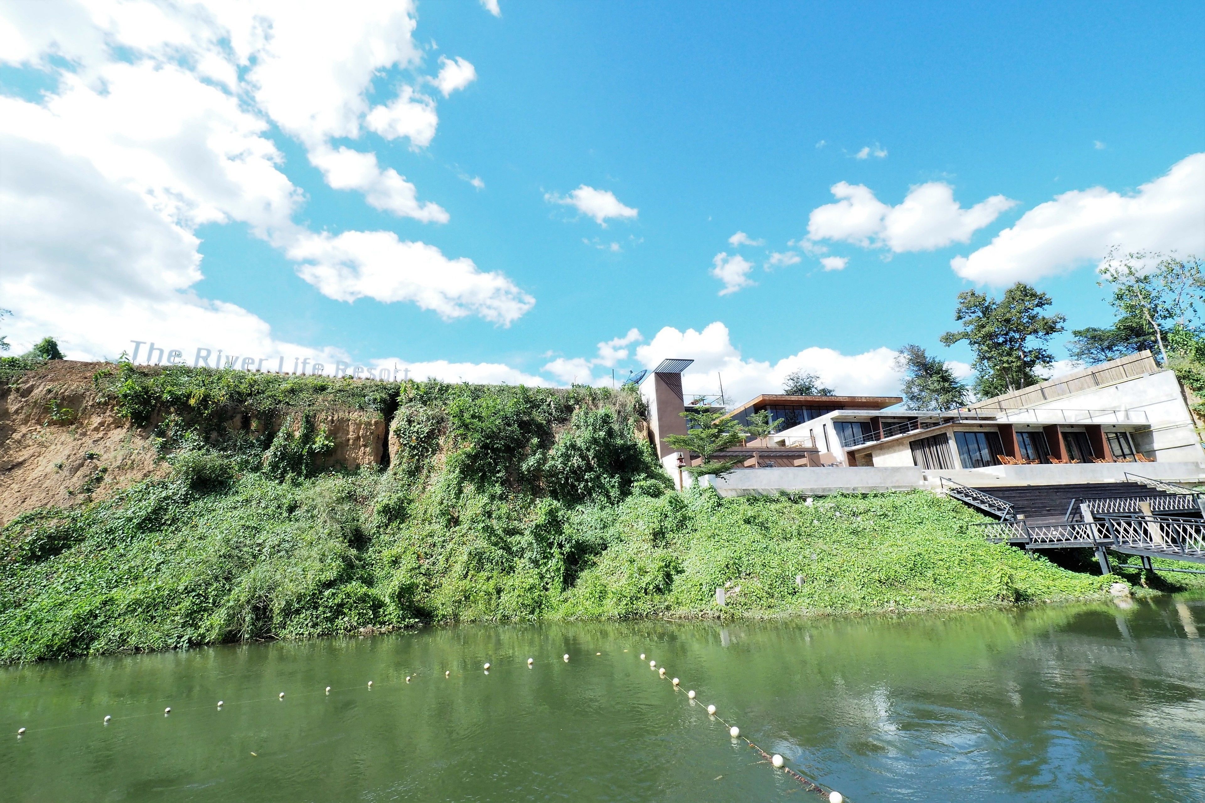 A riverside hotel with a modern design, featuring a bridge and lush greenery.