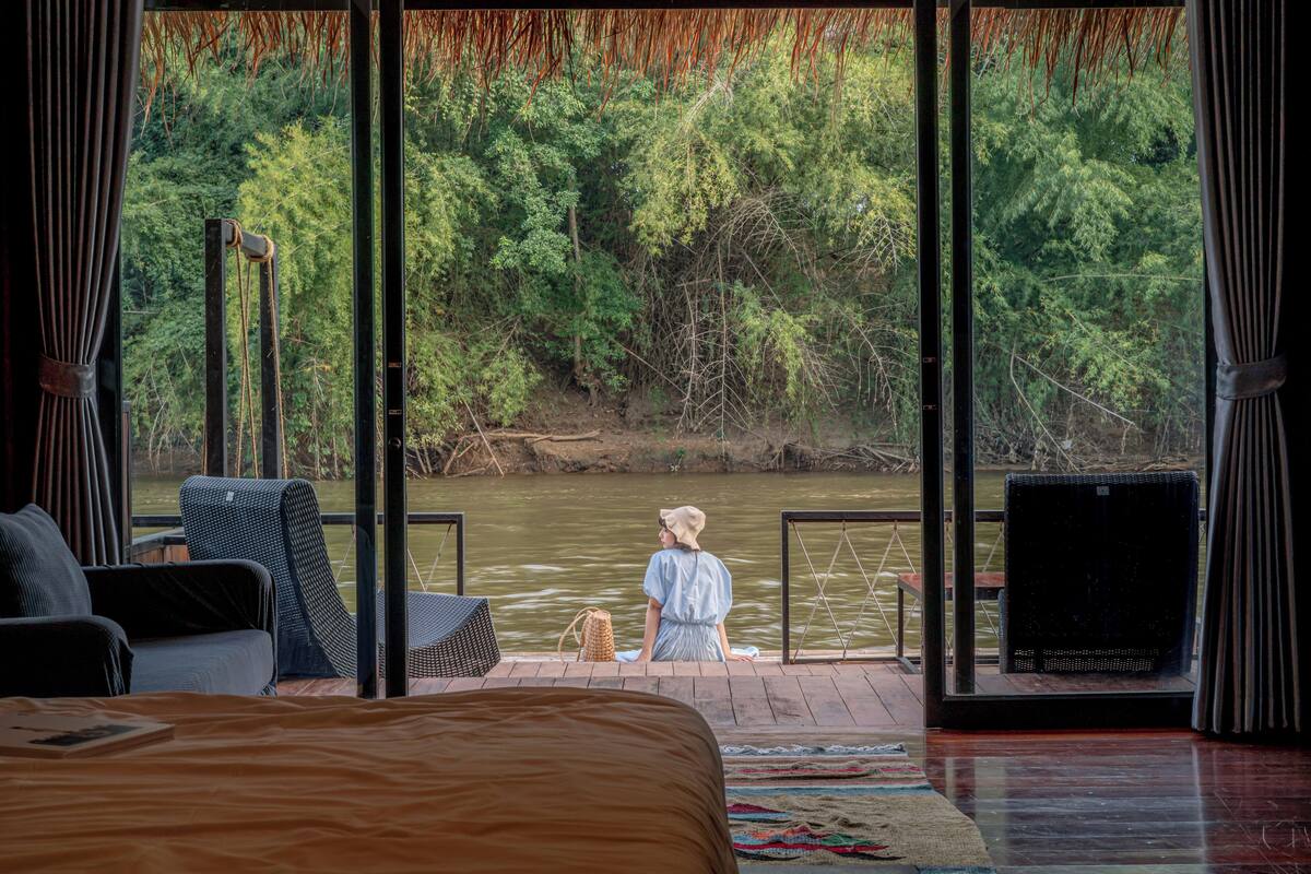 A bedroom with a large window overlooking a river and forest. There is a bed, a chair, and a small table with a book.