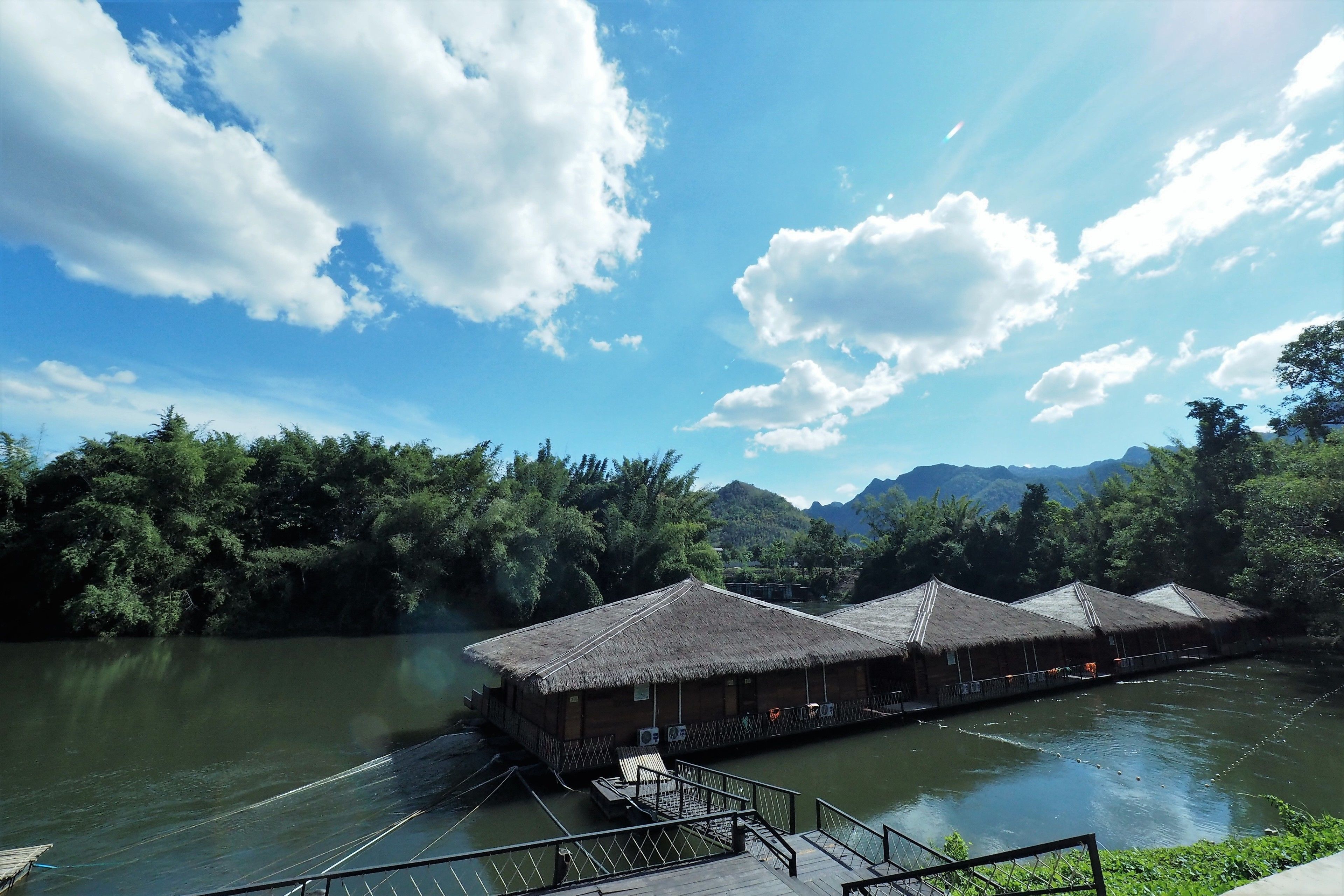 A riverside hotel with thatched-roof structures, a wooden deck, and a view of the river and mountains.