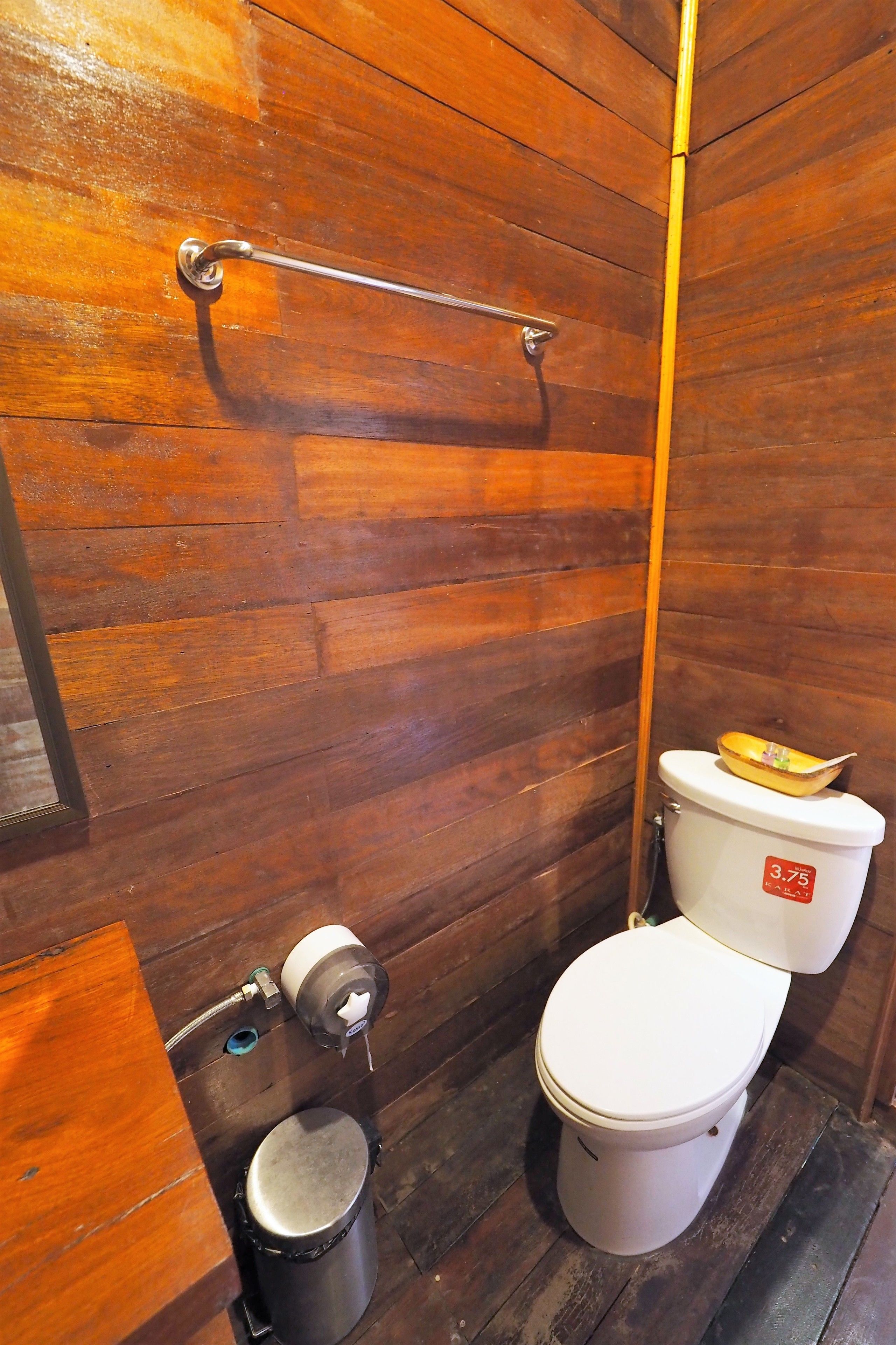 Ein Badezimmer mit Holzwandverkleidung, einer Toilette, einer Metallmülltonne und einem Toilettenpapierhalter.