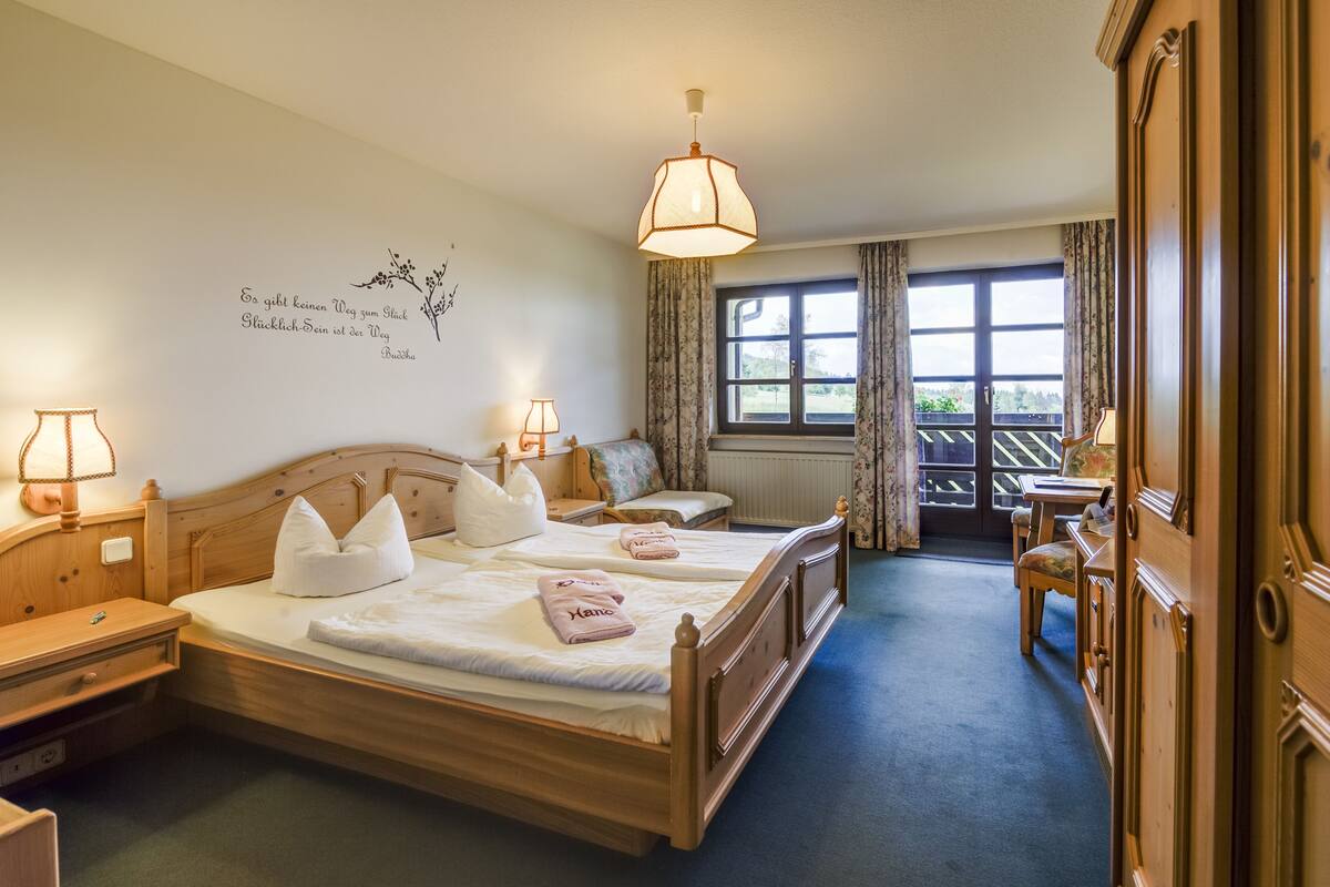 Habitación de hotel con cama de madera, dos mesitas de noche, un escritorio, una silla, ventana con cortinas y vistas al exterior.