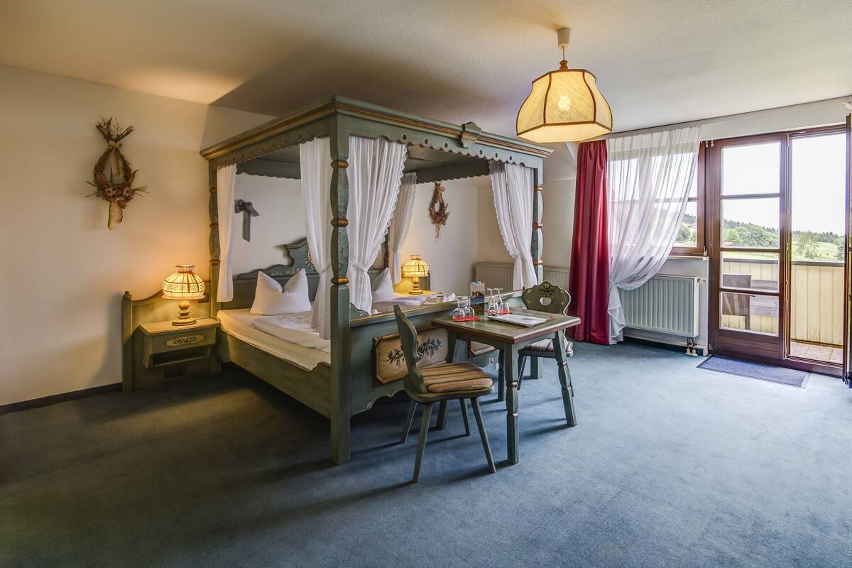 Una cama con dosel y cuatro postes, una mesa con sillas, una lámpara y vistas al exterior a través de una puerta.