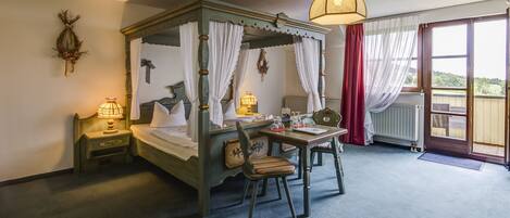 Un lit à baldaquin avec des rideaux blancs, une table avec des chaises, une lampe et une vue sur l’extérieur grâce à une porte-fenêtre coulissante.