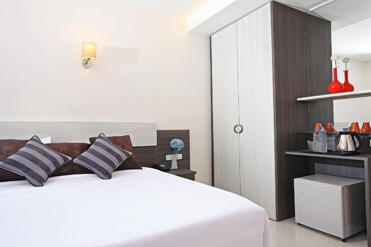 A hotel room with a bed, bedside table, lamp, wardrobe, and a kitchenette area.