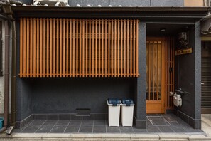 A building with a dark facade, a wooden slatted awning, and two white trash bins outside. - Shiki Homes NENE (Kyoto)