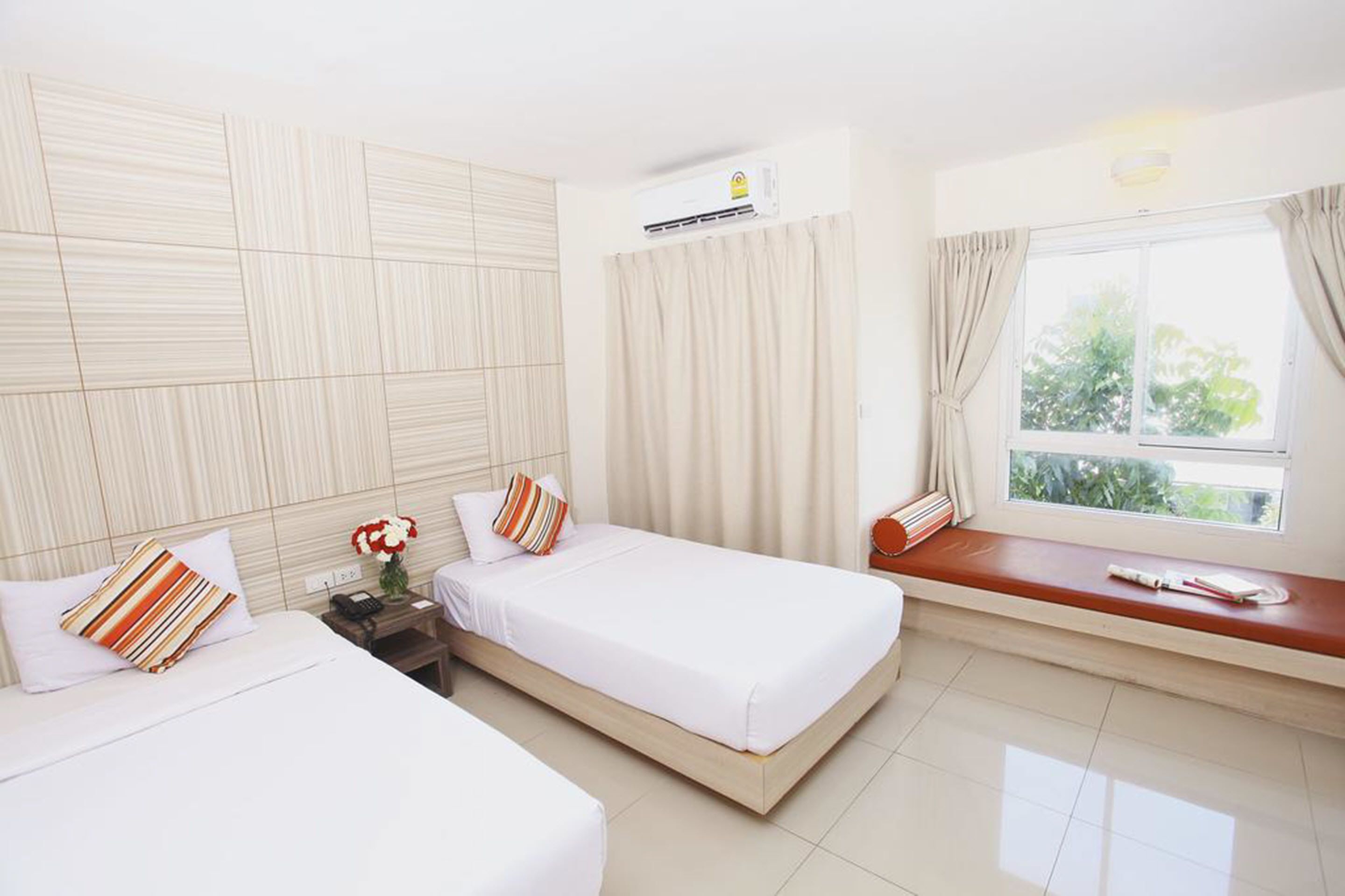 A hotel room with two beds, a window with curtains, an air conditioning unit, and a bedside table with a lamp.