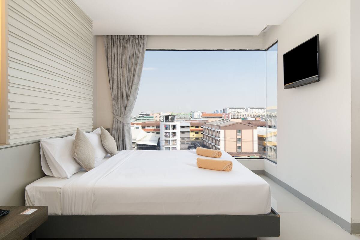 A modern hotel room with a large bed, a flat-screen TV, and a city view through a large window.
