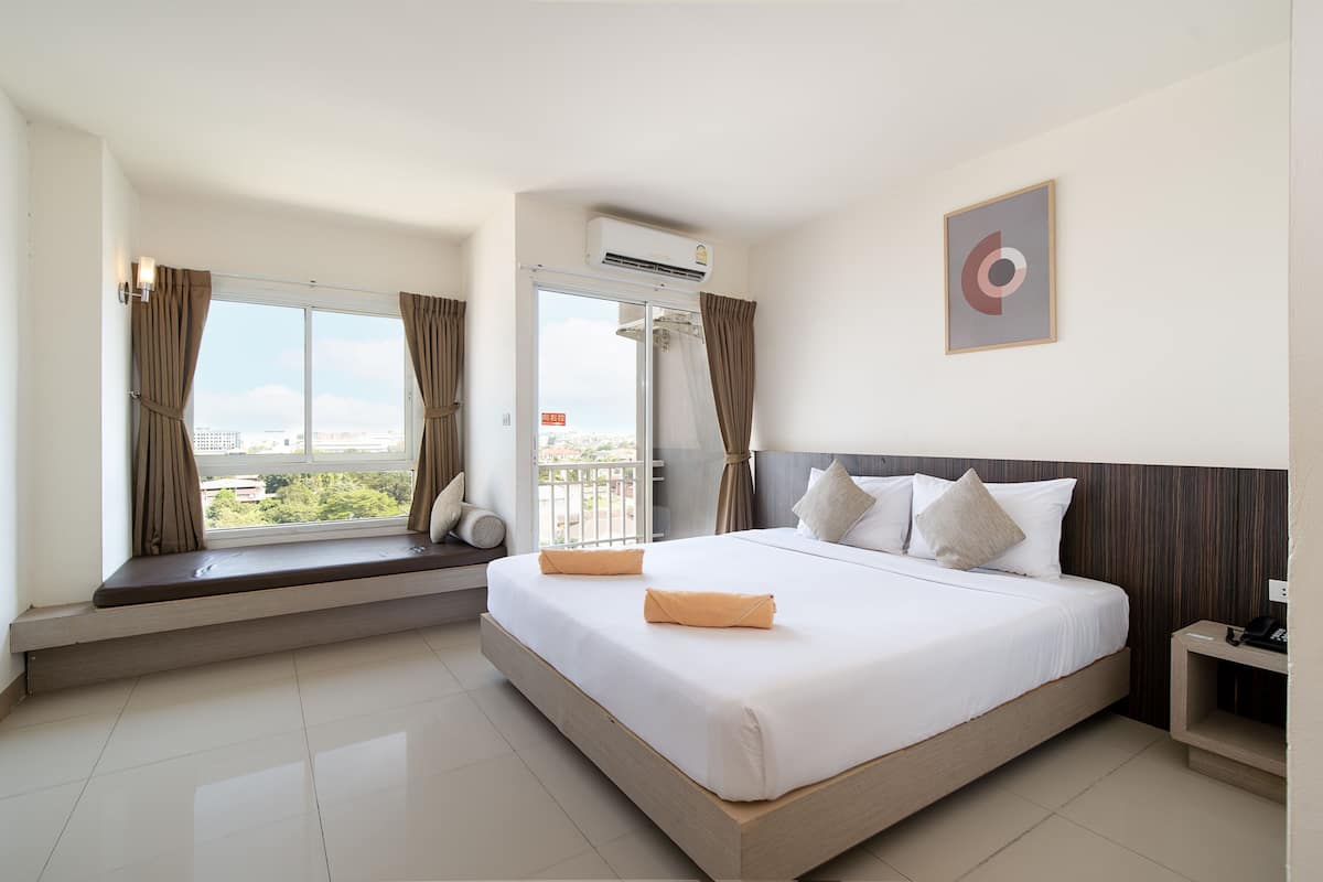 A hotel room with a large bed, a window seat, an air conditioning unit, and a view of the city.