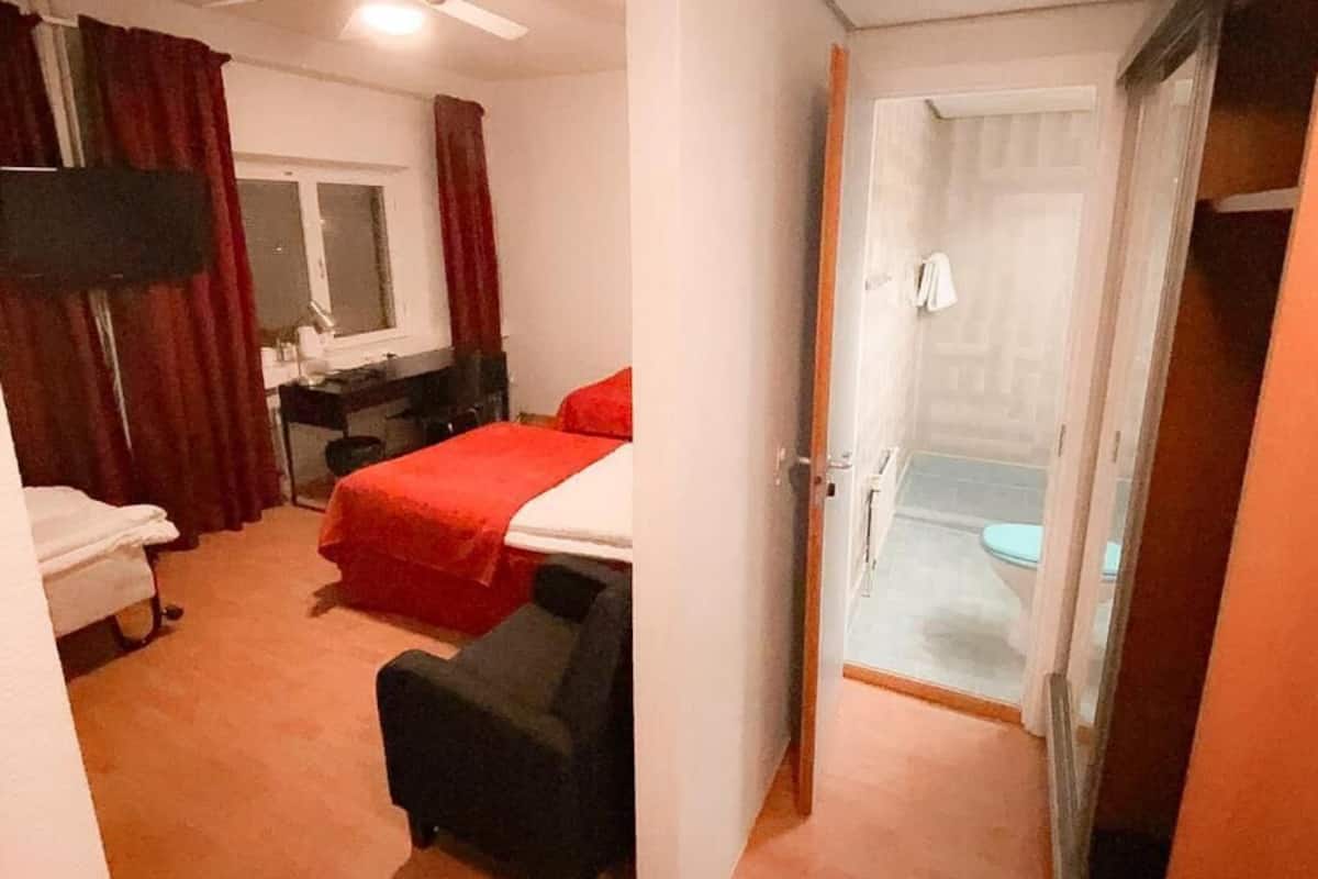A hotel room with a bed, a desk, a chair, a TV, and a bathroom visible through an open door.