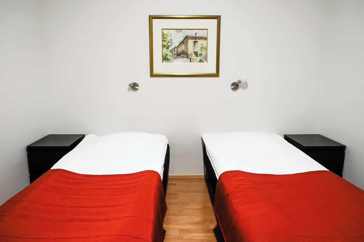 A room with two beds, each with a red bedspread and white pillows, flanked by black nightstands. A framed picture hangs on the wall above the beds.