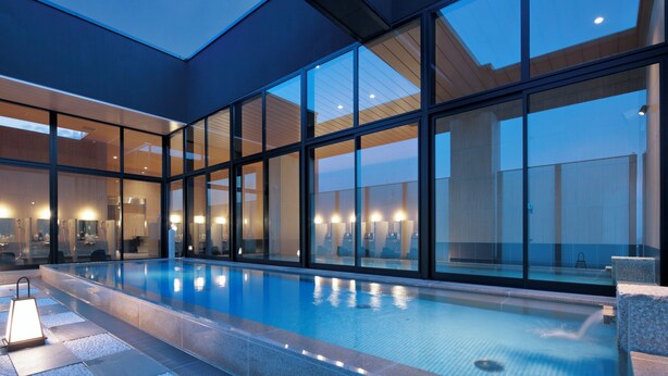 A modern indoor swimming pool with glass walls and a skylight, surrounded by seating areas and a well-lit interior. - Candeo Hotels Osaka Namba (Osaka)