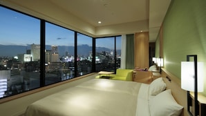 A hotel room with a large bed, a small table, a green armchair, and a view of a cityscape at dusk. - Candeo Hotels Osaka Namba (Osaka)