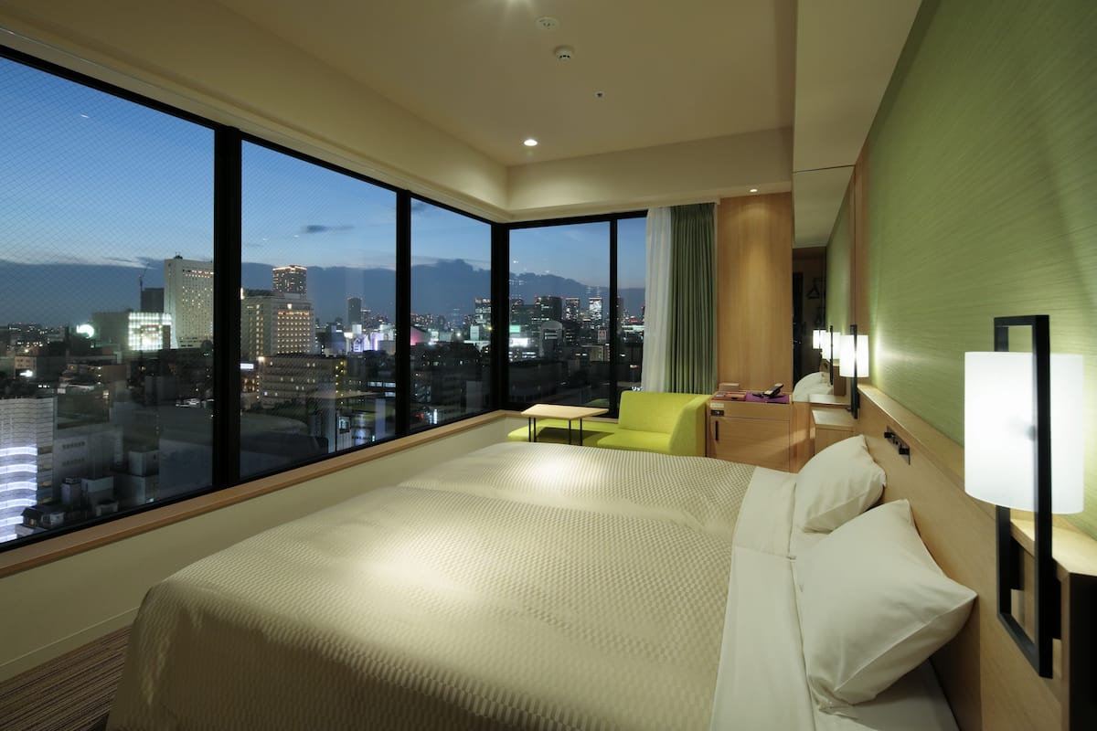 A hotel room with a large bed, a small table, a green armchair, and a view of a cityscape at dusk.