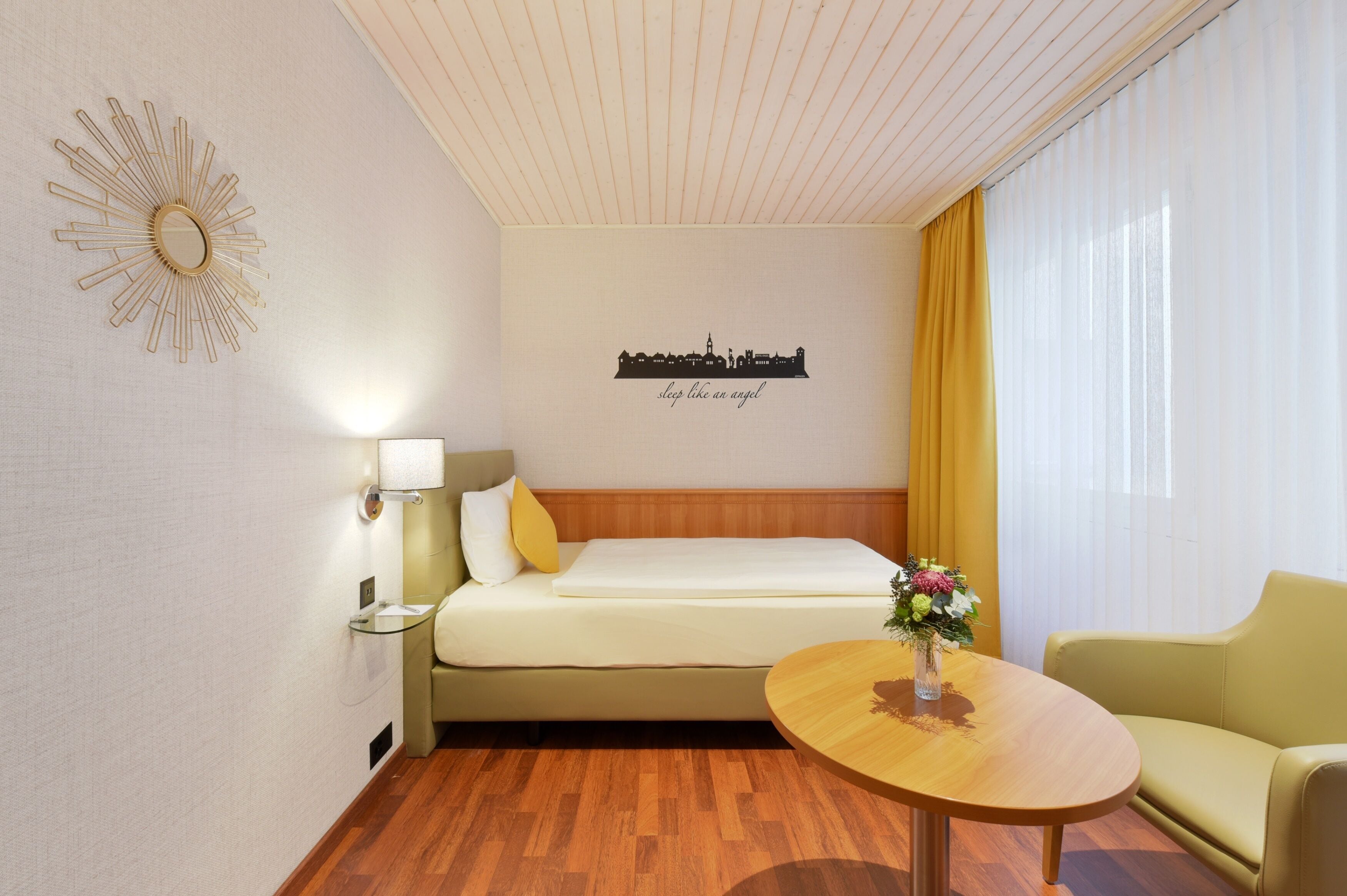 A hotel room with a bed, a sofa, a small table with a vase, a wall-mounted mirror, and a decorative wall piece.
