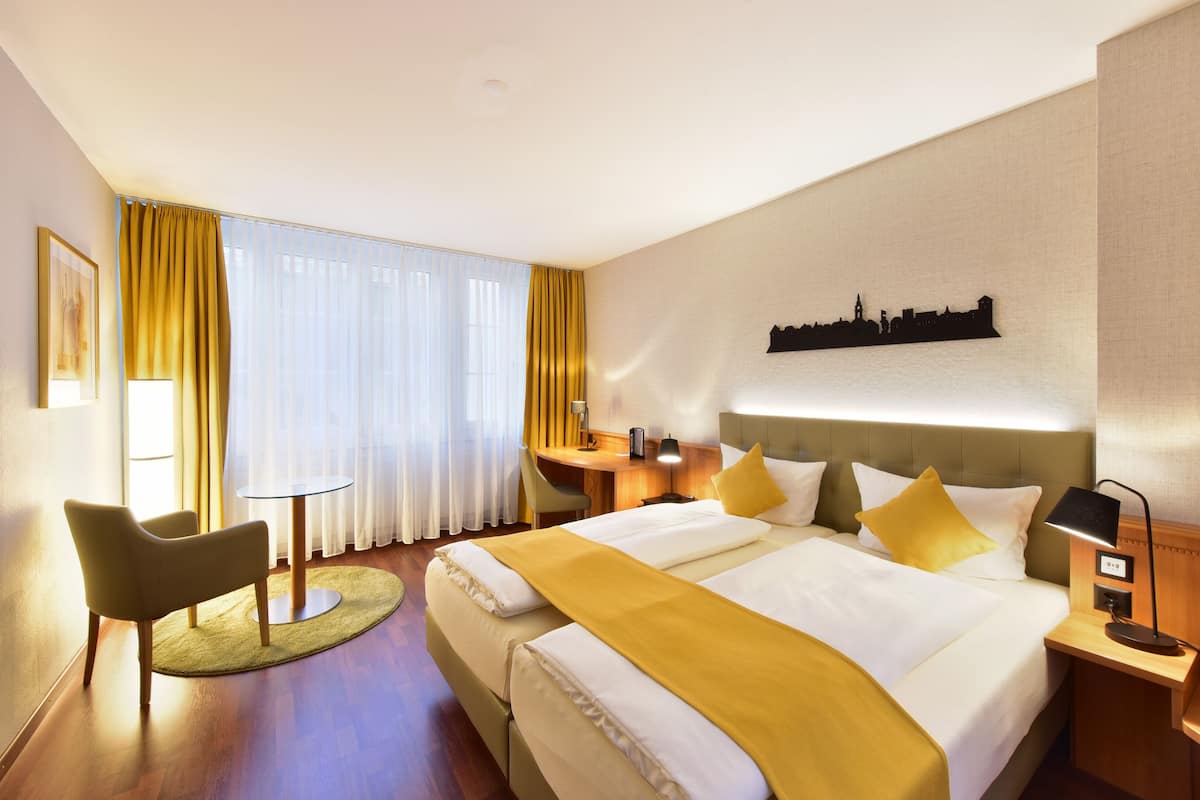 A hotel room with a large bed, a desk, a chair, and a cityscape wall art.
