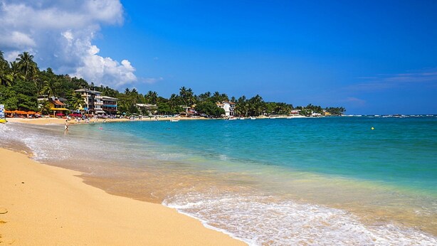 A beach with clear turquoise water, sandy shore, and buildings in the background. - Hotel Happy Night & Restaurant (Unawatuna)