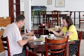 A dining area with wooden chairs and tables, a bookshelf, and a window with a view of greenery. - Hotel Happy Night & Restaurant (Unawatuna)