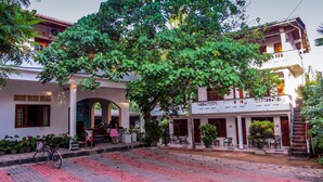 A building with a large tree in front, a paved area with a bicycle, and a staircase leading to a balcony. - Hotel Happy Night & Restaurant (Unawatuna)