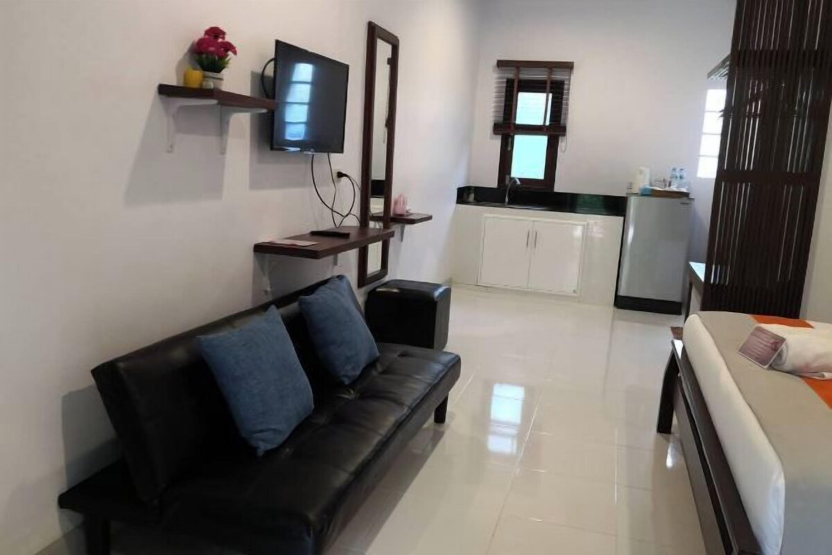 Una sala de estar moderna con un sofá de cuero negro, un televisor de pantalla plana montado en la pared, una pequeña cocina con un refrigerador, y un ventanal que deja entrar luz natural.