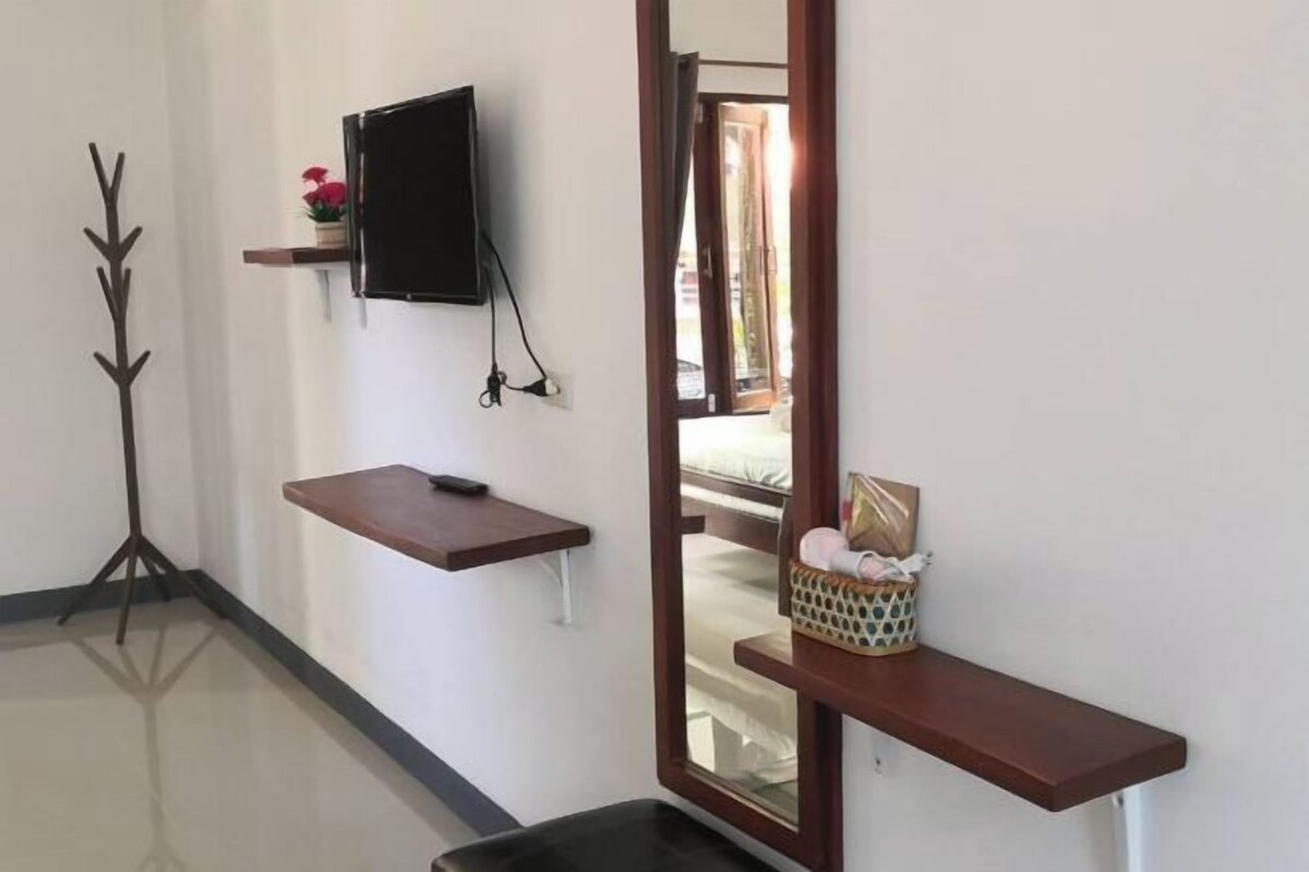 A hotel room with a flat-screen TV mounted on the wall, a small wooden shelf with a remote control, a mirror reflecting the room, and a small basket with items on a wooden shelf.