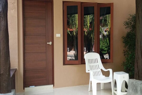 A wooden door, a white plastic chair, and a small stool outside a building with a beige wall and a window with a view of greenery.