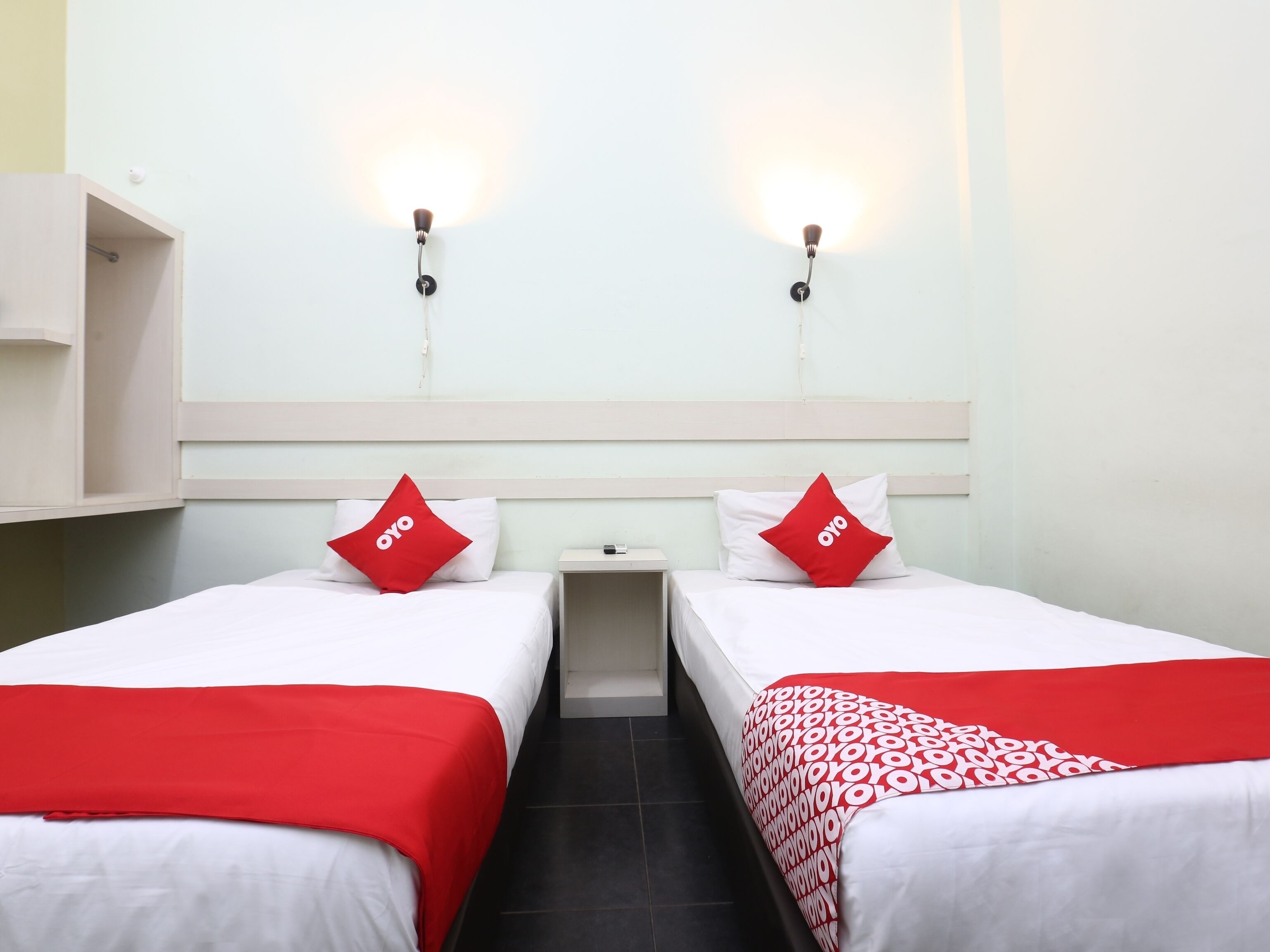 A twin room with two beds, each with a red and white bedspread, a small bedside table, and wall-mounted lights.